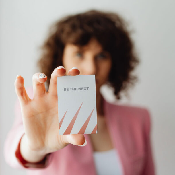 Woman holding a business card with the words "BE THE NEXT" on it.