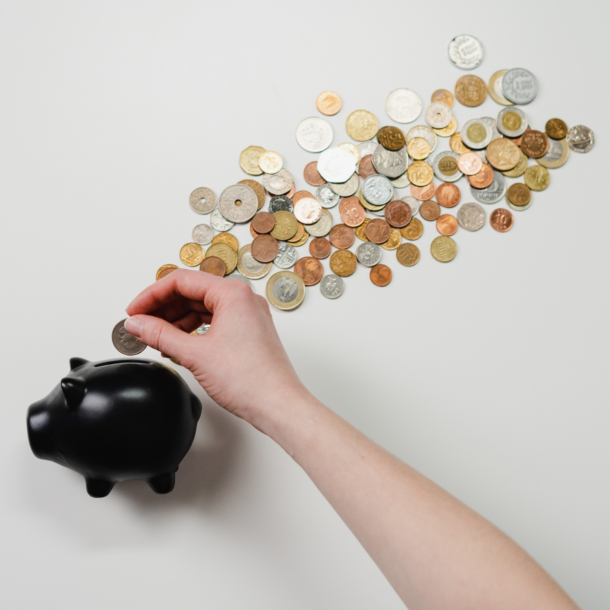 Small black piggy bank with a pile of coins and a Caucasian hand inserting a coin into the bank.