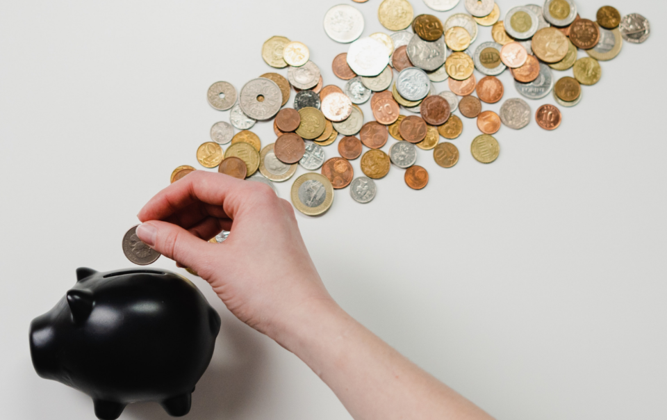 Small black piggy bank with a pile of coins and a Caucasian hand inserting a coin into the bank.