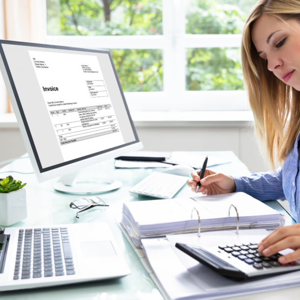 A blonde lady sitting at a desk with a binder, calculator, laptop and monitor in front of her.