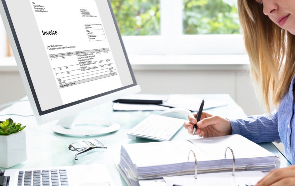 A blonde lady sitting at a desk with a binder, calculator, laptop and monitor in front of her.