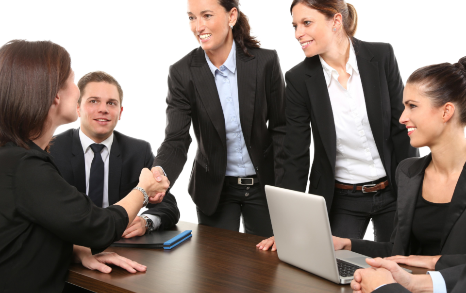 Five colleagues dressed in suits, two are shaking hands, one is on a laptop. They are all sitting at a big wooden table.