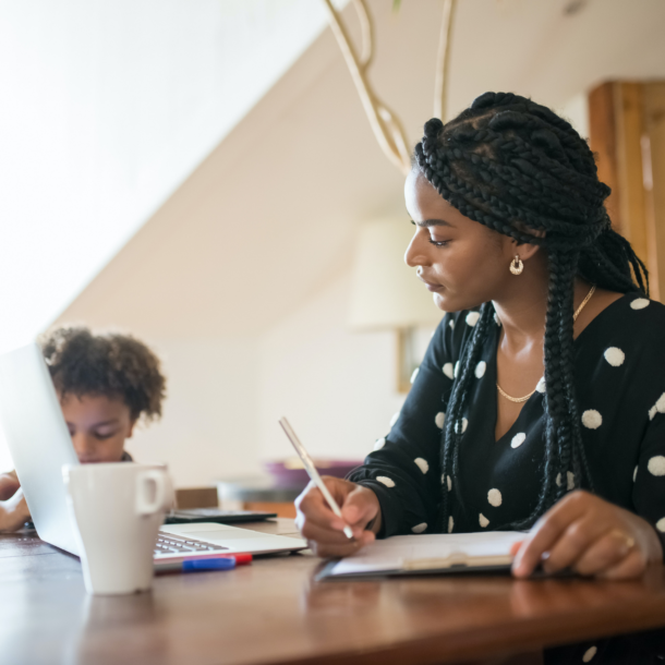 A black woman at home working on her laptop with a child in the background