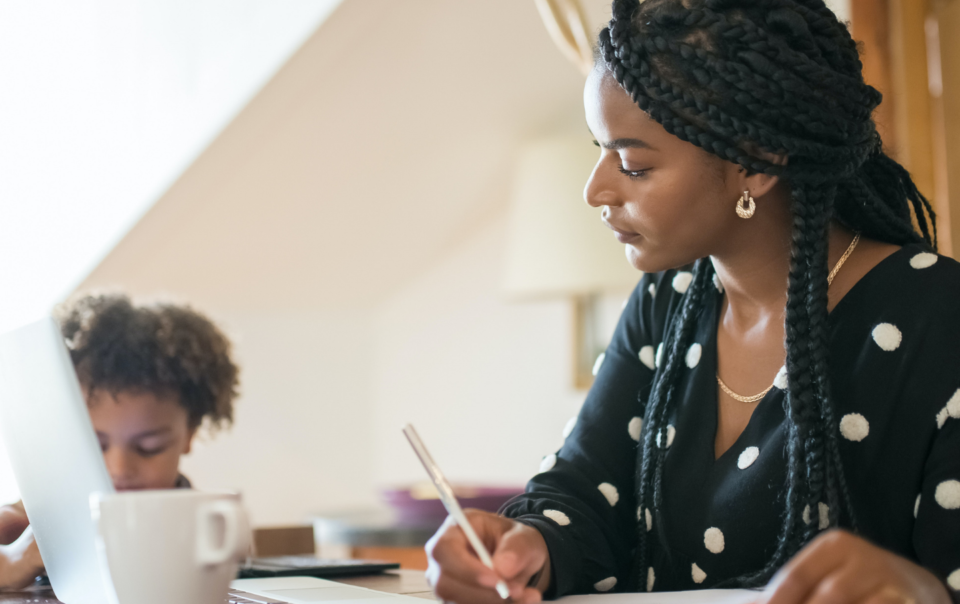 A black woman at home working on her laptop with a child in the background