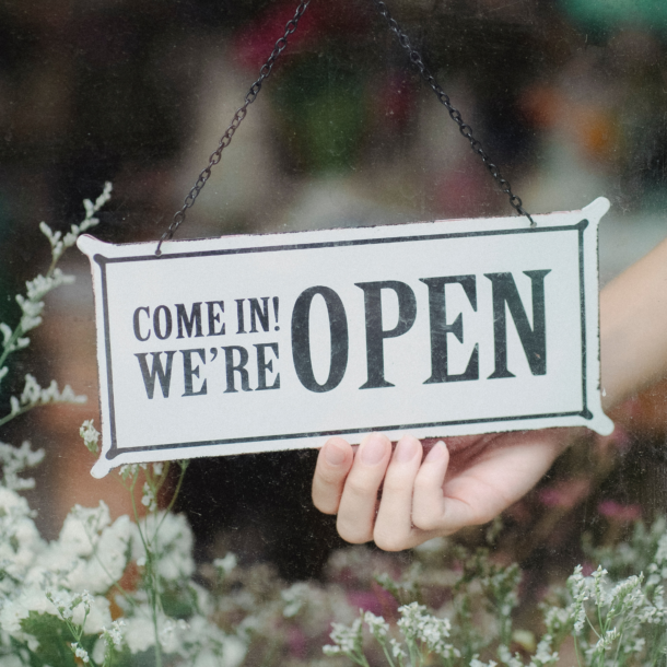 Open sign hanging on a glass door with green leafy plants under the sign.