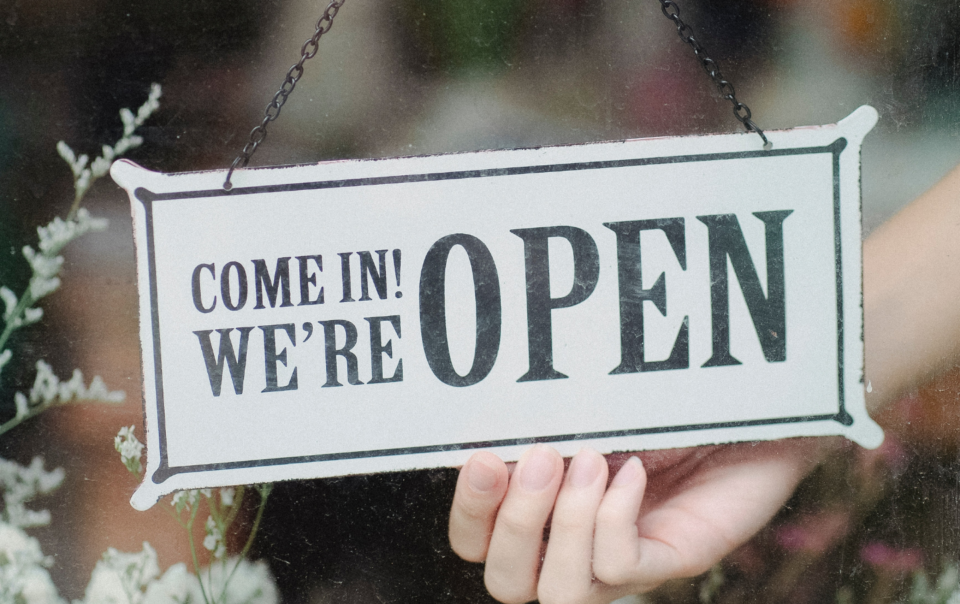 Open sign hanging on a glass door with green leafy plants under the sign.