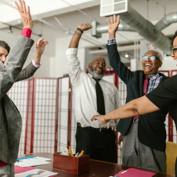 A group of well dressed professionals in a board room standing up and celebrating.